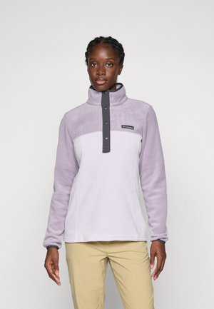 Femme portant un pull en polaire violet clair avec des boutons pression et un pantalon beige, se tenant devant un fond blanc uni.