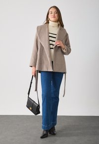 Cappotto in misto lana marrone chiaro con colletto a scialle, abbinato a un dolcevita a righe bianche e nere, jeans blu svasati e stivali neri a punta.