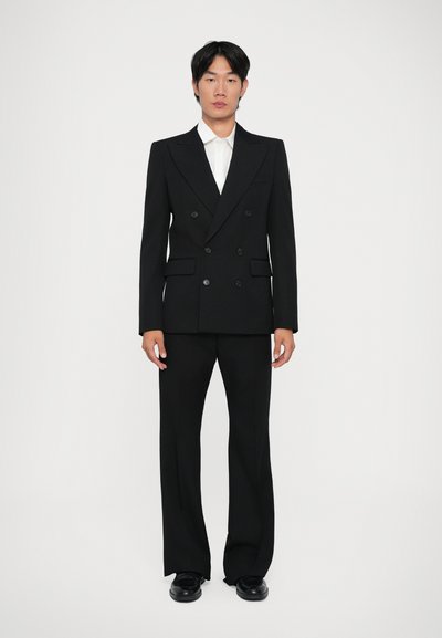 Veste de costume noire à double boutonnage avec larges revers, associée à un pantalon assorti. Comprend une chemise blanche en dessous et des chaussures noires.