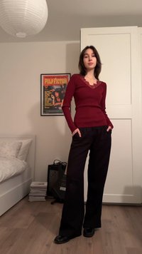 Μπλούζα μπορντό με μακριά μανίκια και δαντελωτή λαιμόκοψη, συνδυασμένη με φαρδιά μαύρη παντελόνια και μαύρα επίπεδα παπούτσια. Το φόντο περιλαμβάνει ένα κρεβάτι και μια αφίσα.