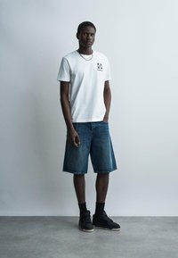 Jeune homme debout contre un mur uni portant un t-shirt blanc avec un motif graphique, un short en jean jusqu’aux genoux, des chaussettes noires et des baskets noires.
