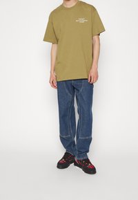 Camiseta de algodón color verde oliva con texto en blanco, combinada con pantalones de mezclilla azul con detalles de costura. Zapatos negros con detalle de cuadros rojos.