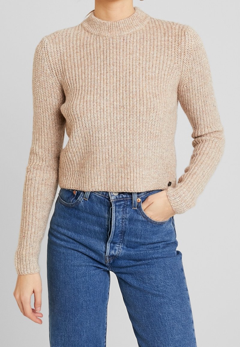 Hellbeige gerippter Pullover mit hohem Kragen und langen Ärmeln, kombiniert mit hochgeschnittenen blauen Jeans. Auffällige Textur und taillierte Passform.