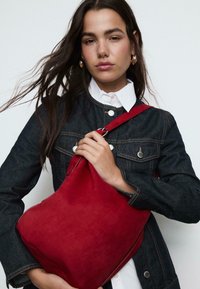 Rdeča semiš torbica z elegantno, zaobljeno obliko in tekstilnim trakom, pritisnjena ob temno modro denim jakno in belo bluzo.