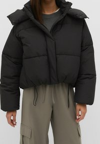 Sort cropped puffer-jakke med oversized ærmer, quiltet tekstur, frontlynlås og trykknapkravedetalje. Sammen med grå bukser.