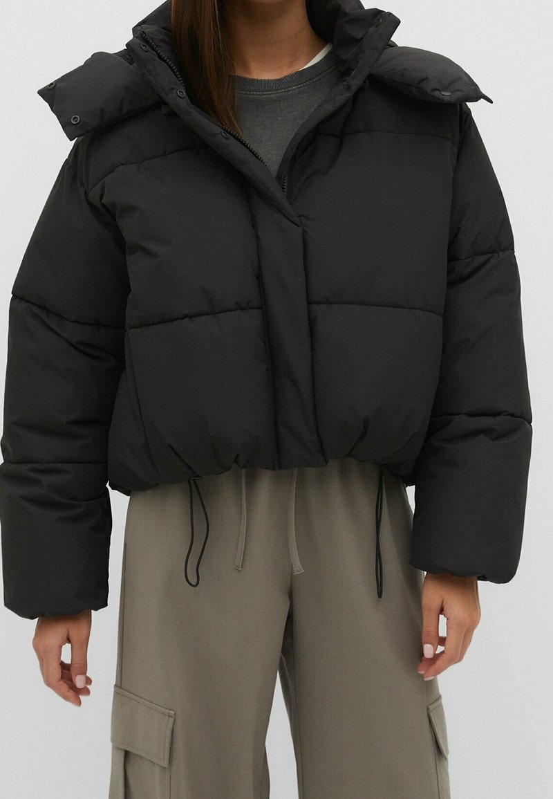 Sort cropped puffer-jakke med oversized ærmer, quiltet tekstur, frontlynlås og trykknapkravedetalje. Sammen med grå bukser.