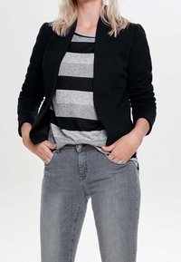 Femme portant une veste noire côtelée sur une chemise rayée grise et noire, les mains dans les poches d'un jean gris ajusté.