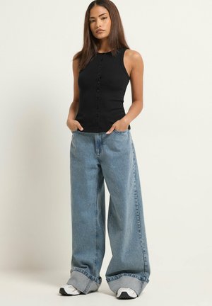 Donna in piedi con le mani nelle tasche, indossa un top nero senza maniche con bottoni, jeans blu a gamba larga con risvolti e sneakers bianche.