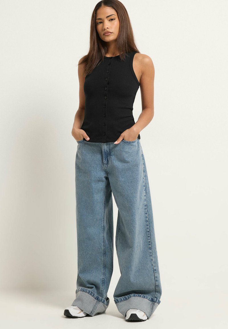 Femme debout, les mains dans les poches, portant un top noir sans manches boutonné, un jean bleu large avec revers et des baskets blanches.