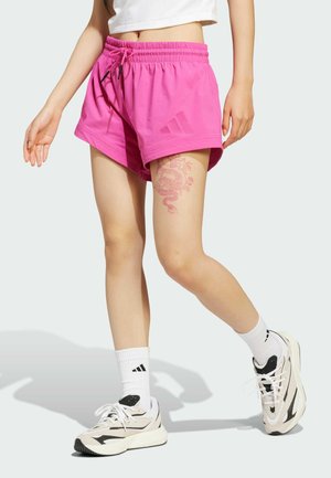 Rosa Sportshorts mit elastischem Bund und Kordelzug. Getragen mit weißen Socken und hellen Schuhen mit schwarzen Akzenten. Sichtbares Drachen-Tattoo.