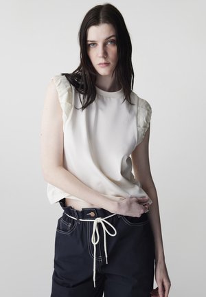 Jeune femme aux longs cheveux noirs porte un débardeur blanc et un pantalon noir avec cordon blanc, posant devant un fond clair uni.