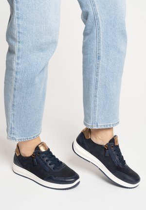 Persona che indossa jeans azzurri e sneakers nere con suole bianche e dettagli bronzo, in piedi su una superficie chiara.