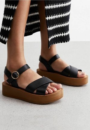 Schwarze Plattform-Sandalen mit überkreuzten Lederriemen und einer runden Schnalle. Strukturierten Riemen stehen im Kontrast zur glatten, sandfarbenen Sohle.