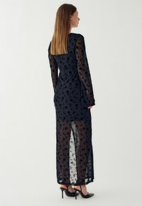 Čierne priesvitné šaty s dlhými rukávmi, kvetinovým vzorom a bočnými rozparkom. Materiál vyzerá byť ľahký a vrstvený. V kombinácii s čiernymi vysokými podpätkami.