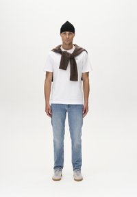 Tricou alb, pulover marron legat în jurul gâtului, blugi deschiși la culoare și adidași bej. Căciulă din lână neagră. Design simplu, casual.