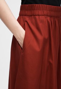 Χέρι που ακουμπά στην τσέπη ενός φαρδιού παντελονιού με ελαστική ζώνη σε σκουριασμένο χρώμα σε απλό φόντο.