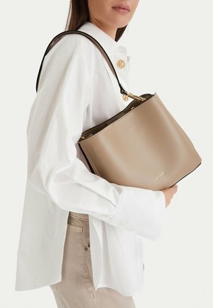 Donna con indosso una camicia bianca e pantaloni beige tiene una borsa tote taupe di Gino Rossi con manici neri sulla spalla.