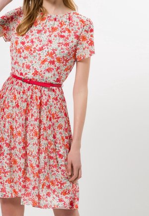 Mujer con vestido blanco de manga corta con estampado floral rojo y rosa y cinturón rojo, de pie sobre un fondo blanco liso.