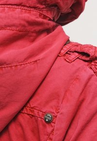 Rode stoffen jas met capuchon, voorzien van zichtbare stiksels en een knoopdetail met het label "KYW 012001." Soepele textuur en getailleerde pasvorm.