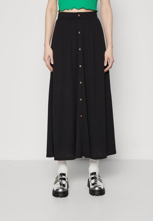 Femme portant une jupe midi noire taille haute boutonnée à l'avant, un top court vert, des chaussettes blanches et des chaussures argentées à grosse boucle.