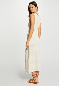 Knieumantel, ärmelloses cremefarbenes Kleid aus strukturiertem Stoff mit geometrischen Mustern; kombiniert mit braunen Sandalen mit Riemchen.