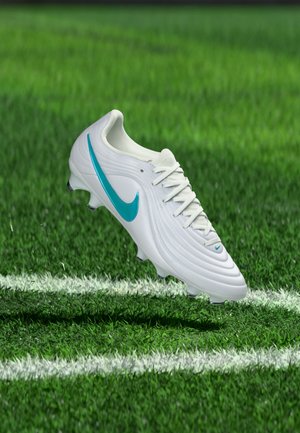 Botín de fútbol blanco con el logo azul de Nike suspendido sobre un campo de césped verde con líneas blancas.