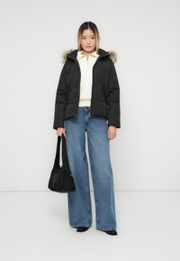 Ung kvinde iført en sort dunjakke med pelskrave, hvid zip-sweater, vide blå jeans, sorte sko, mens hun holder en sort håndtaske.