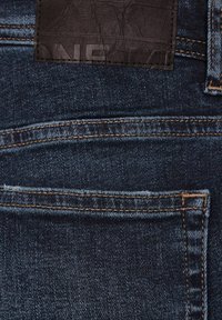 Jeans de mezclilla azul oscuro con un parche de cuero negro texturizado, costuras en contraste y diseño clásico de bolsillos.