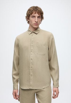 Ung mand med bølget lyst brunt hår iført beige langærmet skjorte med knapper og matchende bukser mod en ensfarvet baggrund.