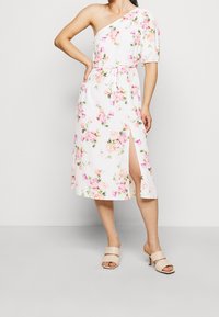 White floral dress featuring one-shoulder design, elastic waist, and side slit. Floral pattern in pink and green. Paired with nude heeled mules.