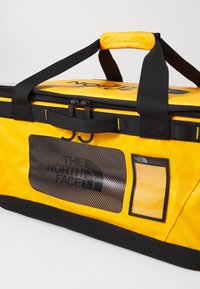 Dzeltena ūdensizturīga duffel soma ar melniem akcentiem, ar rāvējslēdzēju, sietu logu un logotipu. Iespējas ietver izturīgas rokturus un pastiprinātus šuves.