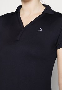 Svart pikétröja med krage, korta ärmar och en liten vit broderad hjärtlogotyp på vänster sida av bröstet. Slät textur.