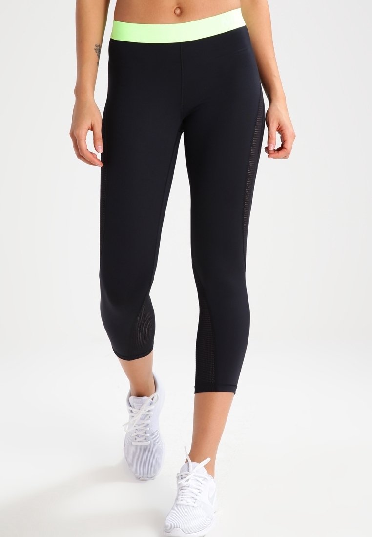 Femme portant des leggings courts noirs avec une ceinture néon verte et des chaussures de sport blanches, debout devant un fond blanc uni.