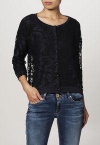 Cardigan in pizzo nero con maniche lunghe, chiusura a bottoni e orlo solido. Indossato sopra un semplice top e abbinato a jeans di denim.