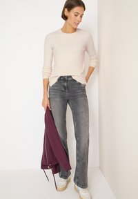 Helles beige gestricktes Sweatshirt, graue Jeans und weiße Sneakers mit blauen Akzenten. Das Modell hält eine burgunderrote Jacke und steht vor einer weißen Wand.