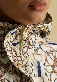 Mūza ar rakstu ūdensnecaurlaidīga jaka ar baltu pamatu, zelta rāvējslēdzēju un aparatūru, kā arī krāsainiem jāšanas motīviem. Redzama faktūru apkakles detaļa.