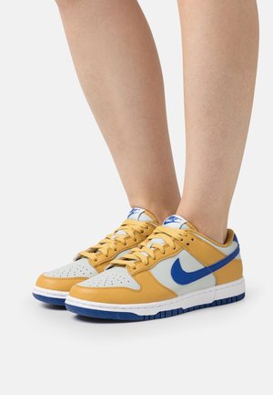 Jambes portant des baskets basses jaunes, gris clair et bleues avec des lacets, un logo Nike bleu et des semelles intermédiaires blanches sur fond uni.
