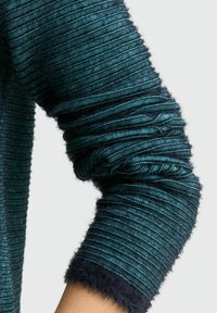 Textured teal sweater sleeve with ribbed pattern and fuzzy black trim at the cuff, showing a blend of materials and a fitted design.