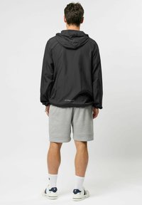 Mees musta kapuutsiga jope, hallide põlvikpükste, valgete sokkide ja valgete tossudega, seisab seljaga vaataja poole tavalise tausta ees.