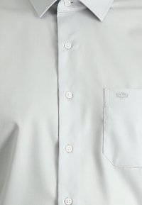 Camicia button-up grigio chiaro con una texture liscia, dotata di collo, sei bottoni e una tasca sul petto con un logo ricamato.