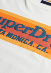 Witte stof met SuperDry-logo gedrukt in blauw op rode en oranje horizontale strepen, inclusief tekst "SANTA MONICA, CA."