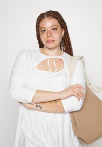 Vestido branco de mangas longas com um detalhe recortado na gola, combinado com uma mala texturizada bege. A modelo tem cabelo vermelho trançado e tatuagens visíveis.