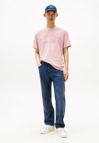 Roze katoenen T-shirt met de opdruk "TOMMY JEANS NEW YORK CITY", gecombineerd met donkerblauwe denim jeans en witte sneakers, aangevuld met een marineblauwe pet.