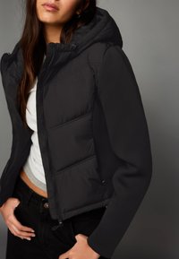 Giacca nera con cappuccio, presenta una parte anteriore trapuntata, tessuto liscio, chiusura con zip e tasche laterali. Indossata sopra una maglia grigia.