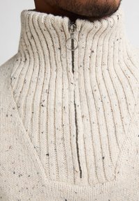 Maglione a maglia color crema con colletto alto a coste e mezza zip. Presenta un motivo testurizzato con toni di colore sparsi.