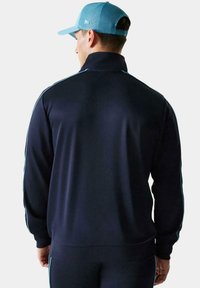 Chaqueta de chándal marina con cuello alto, corte ajustado y franjas laterales en azul claro; textura suave y mangas largas.