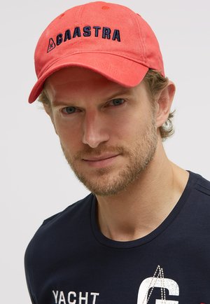 Man with light beard wearing a red Gaastra cap and navy blue shirt with white and red yacht-themed print on a light background.