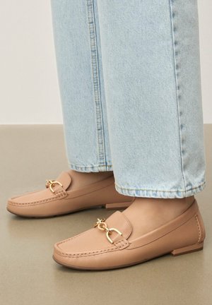 Béžové kožené mokasíny s plochou podrážkou, zlatými kovovými detailmi na priehlavku a viditeľným prešívaním; nosené s svetlomodrými džínsami.