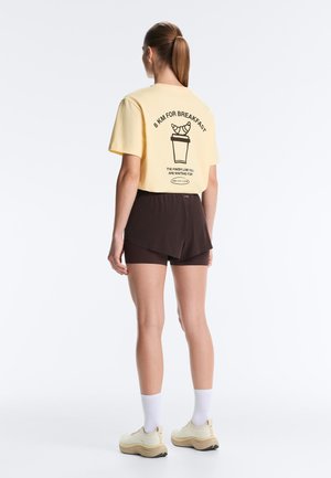 Femme debout, dos visible, portant un t-shirt jaune avec texte noir et illustration de croissant, short noir, chaussettes blanches et baskets beige.