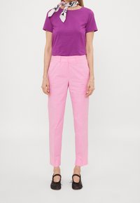 Vrouw in een paarse top met korte mouwen, roze hoge taille broek, zwarte ballerina's en een gestreepte halsdoek tegen een effen achtergrond.